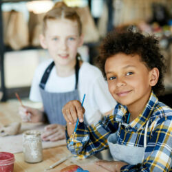 Happy mixed-race boy with paintbrush looking at you at lesson of art and craft